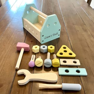 May include: A wooden toy tool box in light blue with the text Liam's Tools printed on the side. The set includes a pink hammer, a light wood wrench, a light blue screwdriver, various colored nuts, bolts, and flat wooden pieces with holes.