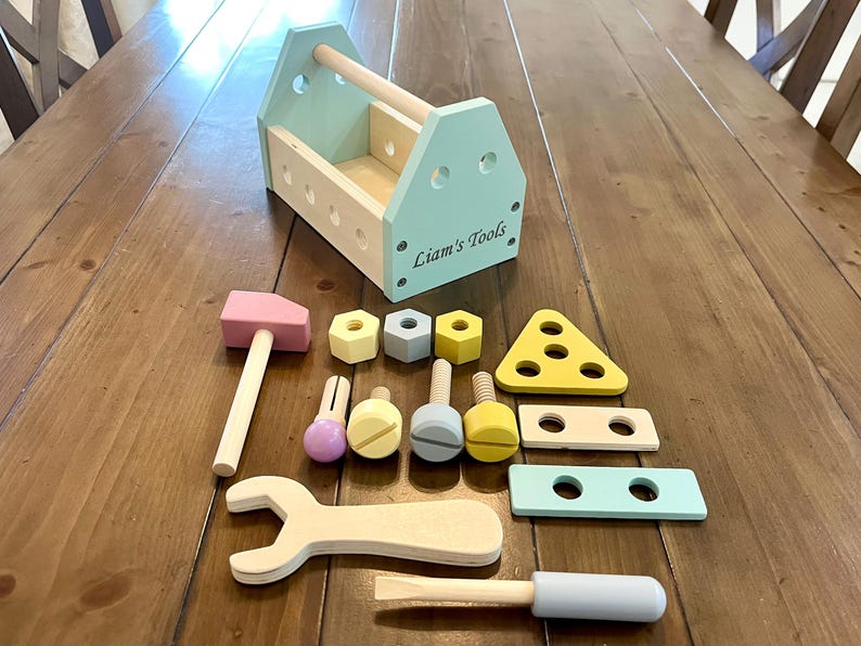 May include: A wooden toy tool box in light blue with the text Liam's Tools printed on the side. The set includes a pink hammer, a light wood wrench, a light blue screwdriver, various colored nuts, bolts, and flat wooden pieces with holes.