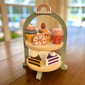 May include: A wooden tiered dessert stand with a rainbow arch and various toy desserts. The desserts include cupcakes, cake rolls, and slices of cake. The stand is painted green and has two wooden shelves.