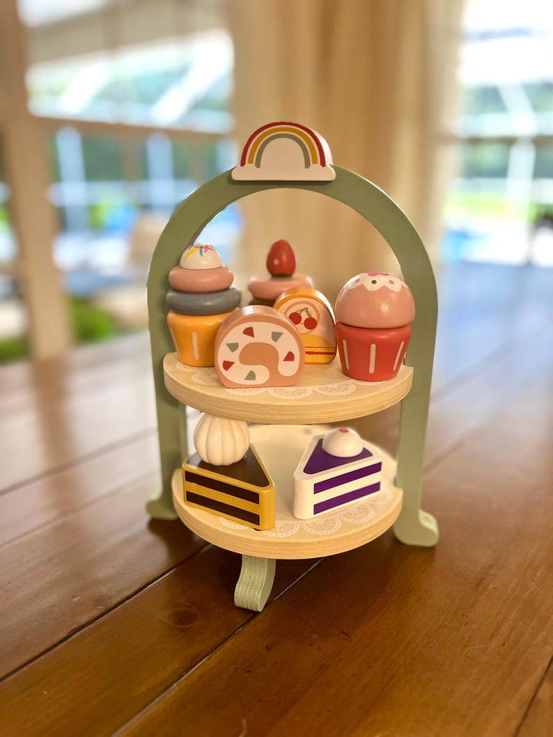 May include: A wooden tiered dessert stand with a rainbow arch and various toy desserts. The desserts include cupcakes, cake rolls, and slices of cake. The stand is painted green and has two wooden shelves.