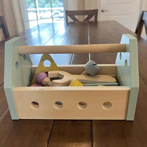 May include: A wooden toy toolbox with light green side panels and a natural wood handle. The box contains various wooden toy tools and components in pastel colors, including a yellow triangle, a grey bolt, and a light purple-topped wooden piece.