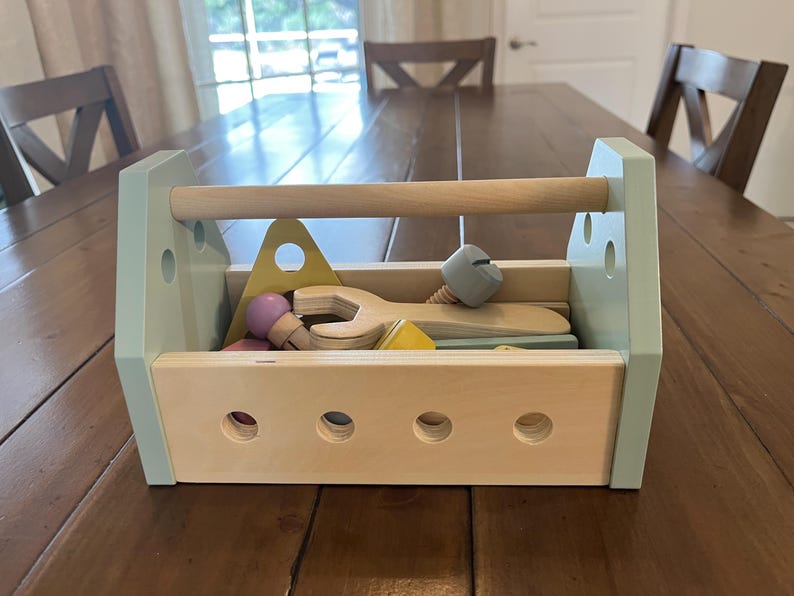 May include: A wooden toy toolbox with light green side panels and a natural wood handle. The box contains various wooden toy tools and components in pastel colors, including a yellow triangle, a grey bolt, and a light purple-topped wooden piece.