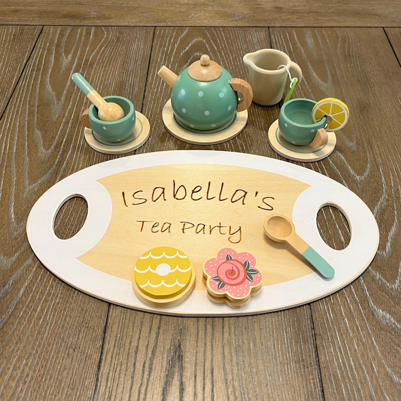 May include: A wooden tea set with a serving tray that reads "Isabella's Tea Party." The set includes a teapot, creamer, cups, saucers, spoons, and decorative treats. The teapot and cups are teal with white polka dots.