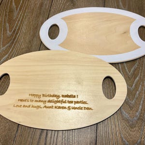 May include: Two wooden serving boards with oval shapes and handle cutouts. One board has a white border. The other board has engraved text that reads: "Happy Birthday, Isabella! Here's to many delightful tea parties. Love and hugs, Aunt Karen & Uncle Dan."