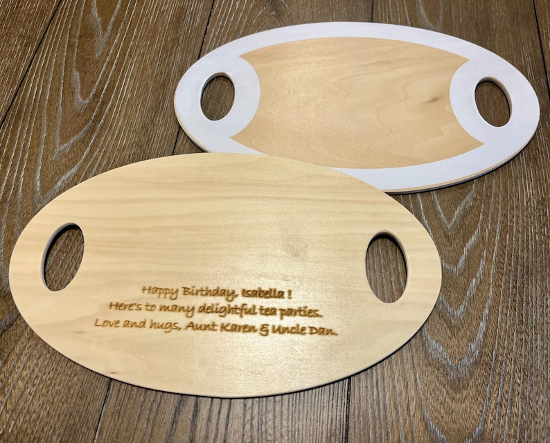 May include: Two wooden serving boards with oval shapes and handle cutouts. One board has a white border. The other board has engraved text that reads: "Happy Birthday, Isabella! Here's to many delightful tea parties. Love and hugs, Aunt Karen & Uncle Dan."