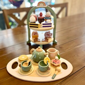May include: Wooden toy tea set with a two-tiered serving tray, a teapot, cups, and desserts. The tea set includes a green teapot with white polka dots, two cups, and a serving tray with a lemon slice and a flower-shaped cookie.