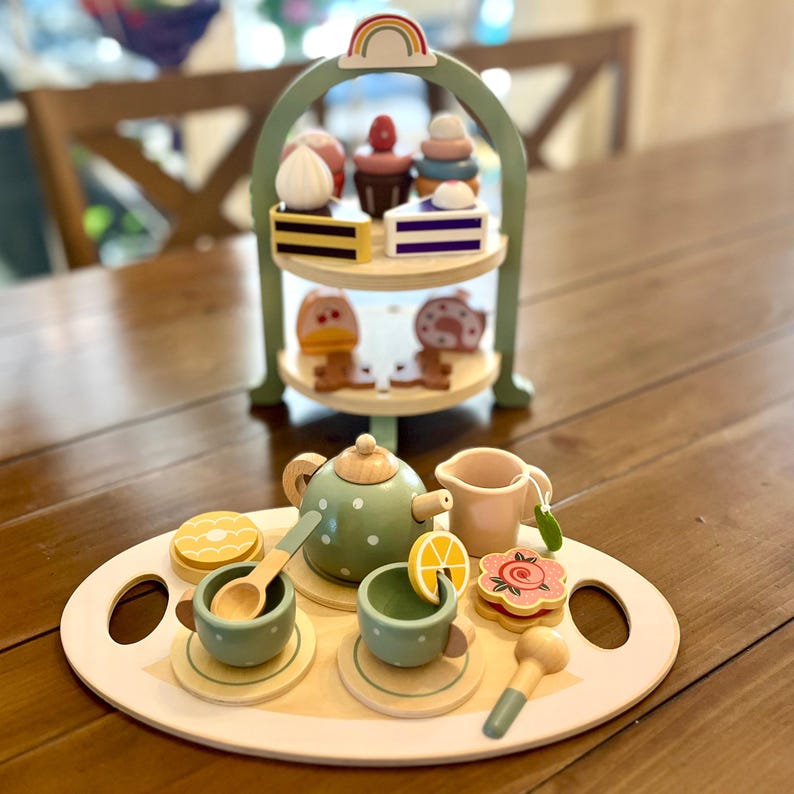 May include: Wooden toy tea set with a two-tiered serving tray, a teapot, cups, and desserts. The tea set includes a green teapot with white polka dots, two cups, and a serving tray with a lemon slice and a flower-shaped cookie.