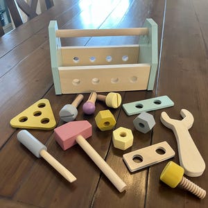 May include: A wooden toy toolbox with a light blue frame and natural wood handle, accompanied by various wooden tools including a hammer, screwdriver, wrench, bolts, nuts, and perforated building plates in pastel yellow, pink, blue, and grey.