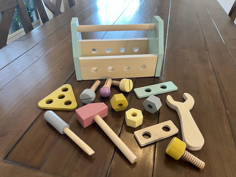 May include: A wooden toy toolbox with a light blue frame and natural wood handle, accompanied by various wooden tools including a hammer, screwdriver, wrench, bolts, nuts, and perforated building plates in pastel yellow, pink, blue, and grey.