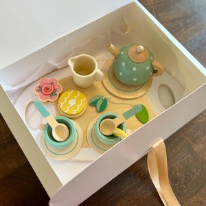May include: A wooden toy tea set in a white box. The set includes a green polka-dot teapot, cups, saucers, spoons, a creamer, and decorative food items. The tea set is designed with pastel colors and natural wood tones, perfect for imaginative play.