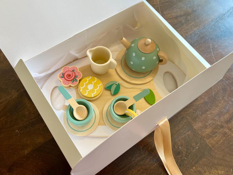 May include: A wooden toy tea set in a white box. The set includes a green polka-dot teapot, cups, saucers, spoons, a creamer, and decorative food items. The tea set is designed with pastel colors and natural wood tones, perfect for imaginative play.