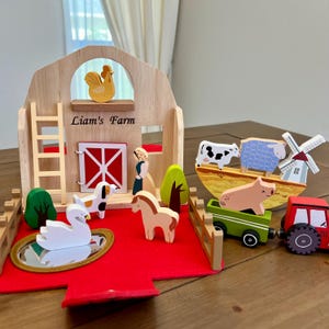 May include: Wooden farm playset featuring a barn with the text "Liam's Farm", a red roof, and various farm animals. Includes a tractor, a pig in a green cart, a cow, a sheep, a windmill, a swan, a horse, and a dog.