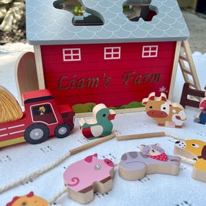 May include: A red wooden barn with a white sign that says "Liam's Farm". The barn has a ladder on the side and a hay bale, tractor, cow, duck, pig, cat, dog, and farmer figurine in front of it.