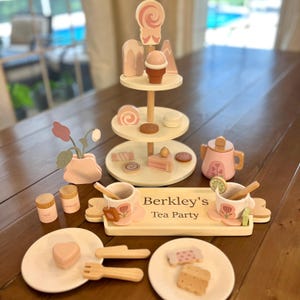 May include: Wooden tea party set with a tiered dessert stand, teapot, cups, plates, and serving tray. The set includes miniature desserts, sugar containers, and a floral arrangement. The tray reads "Berkley's Tea Party".