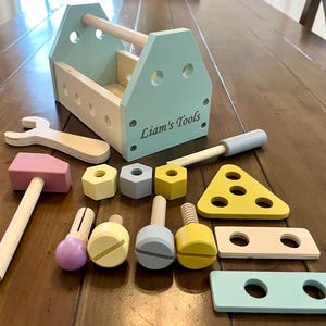 May include: A wooden toy tool set featuring a light blue toolbox with the text Liam's Tools, a pink hammer, a wrench, three hexagonal nuts in yellow and gray, three bolts, a yellow triangular piece, and two rectangular wooden planks with holes.
