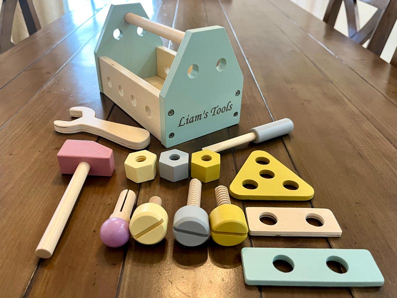May include: A wooden toy tool set featuring a light blue toolbox with the text Liam's Tools, a pink hammer, a wrench, three hexagonal nuts in yellow and gray, three bolts, a yellow triangular piece, and two rectangular wooden planks with holes.