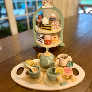 May include: Wooden toy tea set with a two-tiered serving tray, teapot, cups, and desserts. The set includes a light green serving stand with a rainbow on top. The tea set includes a teapot with white polka dots, cups, saucers, and wooden desserts.