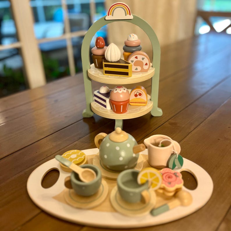 May include: Wooden toy tea set with a two-tiered serving tray, teapot, cups, and desserts. The set includes a light green serving stand with a rainbow on top. The tea set includes a teapot with white polka dots, cups, saucers, and wooden desserts.