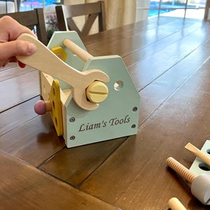 May include: A light blue wooden toy toolbox with the text Liam's Tools printed on the side. The set includes a light wood wrench, a yellow screw, a purple peg, and additional wooden construction pieces with holes and screws in various colors.