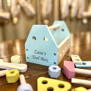 May include: A light blue wooden toy tool box with a natural wood handle and the text Zane's Tool Box printed on the front. The set includes various wooden toy tools and components in pastel colors like yellow, pink, and light blue scattered around.