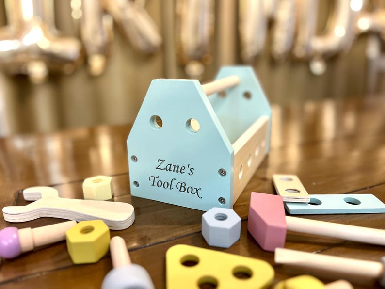 May include: A light blue wooden toy tool box with a natural wood handle and the text Zane's Tool Box printed on the front. The set includes various wooden toy tools and components in pastel colors like yellow, pink, and light blue scattered around.