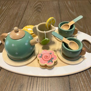 May include: A wooden toy tea set on a white and wood-toned tray. The set includes a green teapot with white polka dots, two cups with spoons, a creamer, a flower-shaped cookie, and a lemon slice.