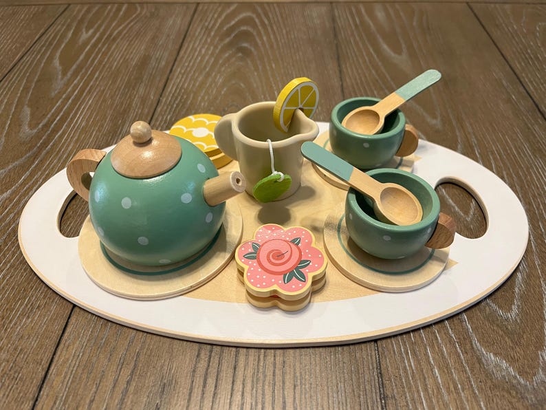 May include: A wooden toy tea set on a white and wood-toned tray. The set includes a green teapot with white polka dots, two cups with spoons, a creamer, a flower-shaped cookie, and a lemon slice.