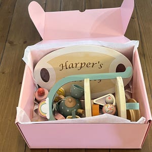 May include: A pink gift box filled with wooden toys, including a tea set, a swing set, and a sign that reads "Harper's". The toys are in various colors, including light blue, orange, and natural wood tones. The box is lined with white tissue paper.