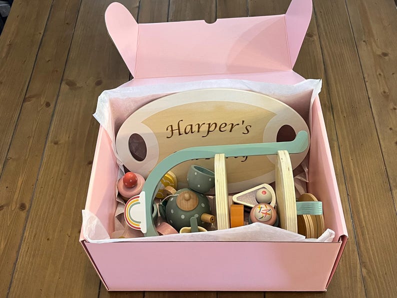 May include: A pink gift box filled with wooden toys, including a tea set, a swing set, and a sign that reads "Harper's". The toys are in various colors, including light blue, orange, and natural wood tones. The box is lined with white tissue paper.