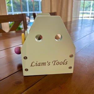 May include: A light green wooden toy tool box with a house-shaped profile, featuring two circular cutouts near the top and the text "Liam's Tools" printed in dark lettering on the front. The structure is secured with four visible metal screws.