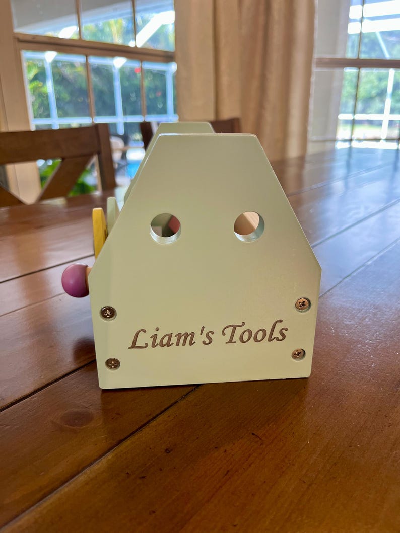 May include: A light green wooden toy tool box with a house-shaped profile, featuring two circular cutouts near the top and the text "Liam's Tools" printed in dark lettering on the front. The structure is secured with four visible metal screws.