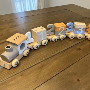 May include: A wooden toy train with a personalized engine that says "Jack" and colorful number blocks. The train is made of natural wood and has a gray engine, white wheels, and a light brown body. The number blocks are stacked on top of each other and are painted in different colors, including orange, blue, and gray.