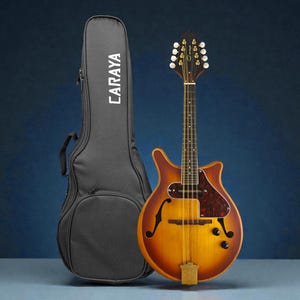 Puede incluir: Mandolina Caraya con acabado sunburst, golpeador marrón oscuro y puente de madera. Una funda negra con el nombre de la marca "CARAYA" está a la izquierda. El instrumento tiene un cuerpo en forma de pera y agujeros en forma de f.