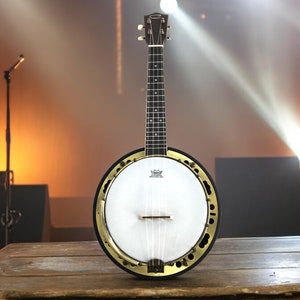 May include: A black and white banjo ukulele with a gold-colored rim and tuning pegs. The banjo ukulele is on a wooden table with a microphone stand and a spotlight in the background.
