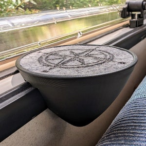 May include: A black, 3D printed cup holder with a grey, compass-shaped insert. The cup holder is mounted to a window frame.