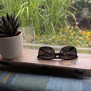 Könnte beinhalten: Eine kleine, dunkelgrüne Sukkulente in einem weißen, gerippten Keramiktopf steht auf einem Holzregal. Schwarze Sonnenbrille liegt auf dem Regal. Das Regal ist an einem Fenster mit Blick auf Grün befestigt.
