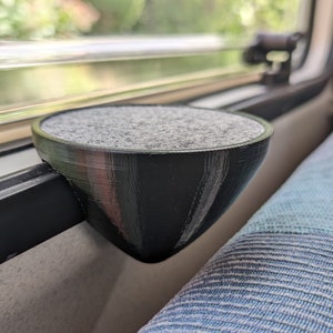 Könnte beinhalten: Ein schwarzer 3D-gedruckter Becher mit grauer Filz-Auskleidung, montiert an einem Fensterrahmen. Der Becher ist so konzipiert, dass er kleine Gegenstände wie Schlüssel oder Münzen aufnehmen kann.