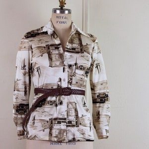May include: A white long-sleeved shirt with a brown and white print of houses and boats. The shirt has a brown woven belt.