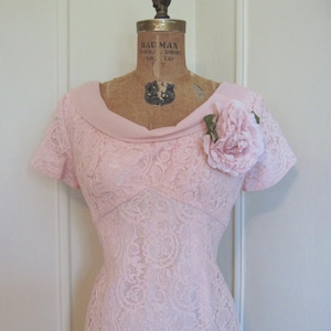 May include: A pink lace dress with a scoop neck and short sleeves. The dress has a fitted bodice and a full skirt. A large pink flower is pinned to the bodice.