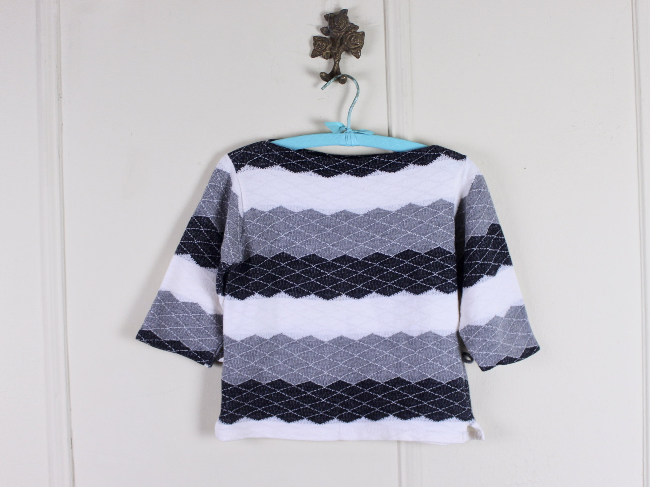 May include: A boat-neck sweater with a geometric pattern in white, grey, and black hangs on a blue hanger. The sweater has three-quarter length sleeves. The pattern consists of horizontal rows of diamond shapes.
