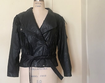 May include: Black leather jacket with fringe detailing on the sleeves and chest. The jacket features a wrap-style closure with a belt and a notched collar. The jacket is displayed on a mannequin.