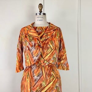 Op de afbeelding: Een vintage jurk en jasset in een abstracte print in oranje, bruin en groen. De jurk heeft een getailleerd bovenstuk en een volle rok. De jas heeft een kraag en driekwart mouwen.