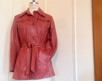 Può includere: Una giacca di pelle rossa con cintura, due grandi tasche e chiusura con bottoni.