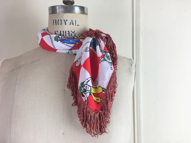 May include: A red and white checkered scarf with a fringe border. The scarf features a colorful cartoon design of people riding horses. The scarf is tied around a white mannequin's neck. The mannequin has the words "ROYAL FORM" printed on the top of its head.