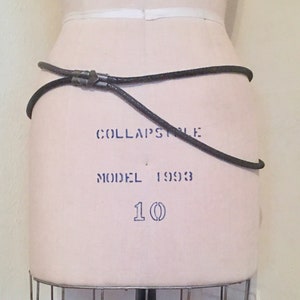 Puede incluir: Un cinturón de cuero negro con una hebilla plateada. El cinturón está en un maniquí blanco. El maniquí tiene el texto "COLLAPSIBLE MODEL 1993 10" impreso en él.