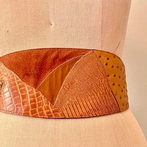 Op de afbeelding: Een brede bruine leren riem met een patchwork-ontwerp. De riem is gemaakt van verschillende soorten leer, waaronder krokodil, struisvogel en suède.