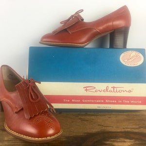 May include: A pair of red leather heeled shoes with fringe detailing and a brogue design. The shoes are sitting on top of a blue and white box that says "Revelations, The Most Comfortable Shoes in The World".