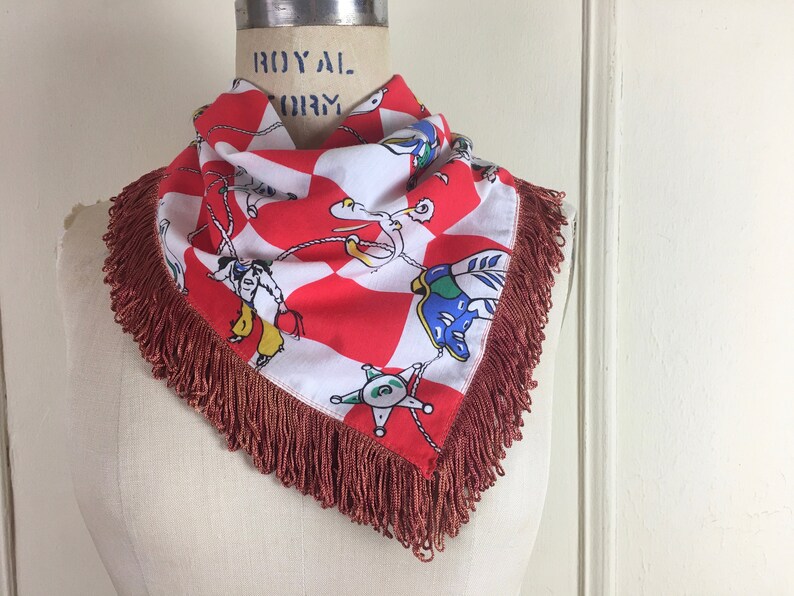 May include: A red and white bandana with a checkered pattern and a fringe border. The bandana features a colorful illustration of cowboys roping a calf.