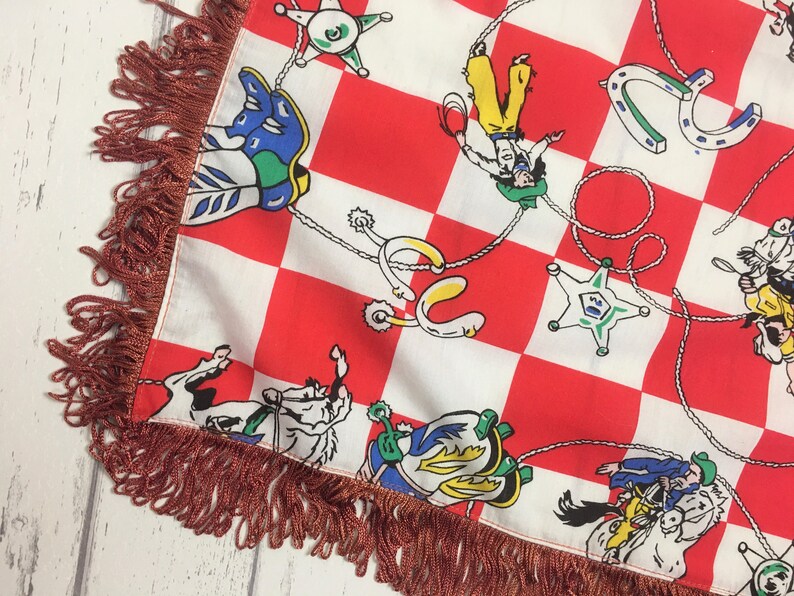 May include: Red and white checkered fabric with a fringe border. The fabric features a repeating pattern of cowboys, horses, and horseshoes.