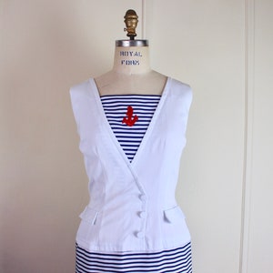 May include: A white sleeveless vest with a button closure over a blue and white striped dress. The dress has a red anchor applique on the front.